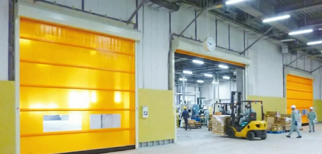 Monban high speed door sedang beroperasi di area pabrik yang sibuk. Dengan dua pintu pabrik otomatis, yang disebelah kanan kondisinya terbuka dan sebelah kiri tertutup.